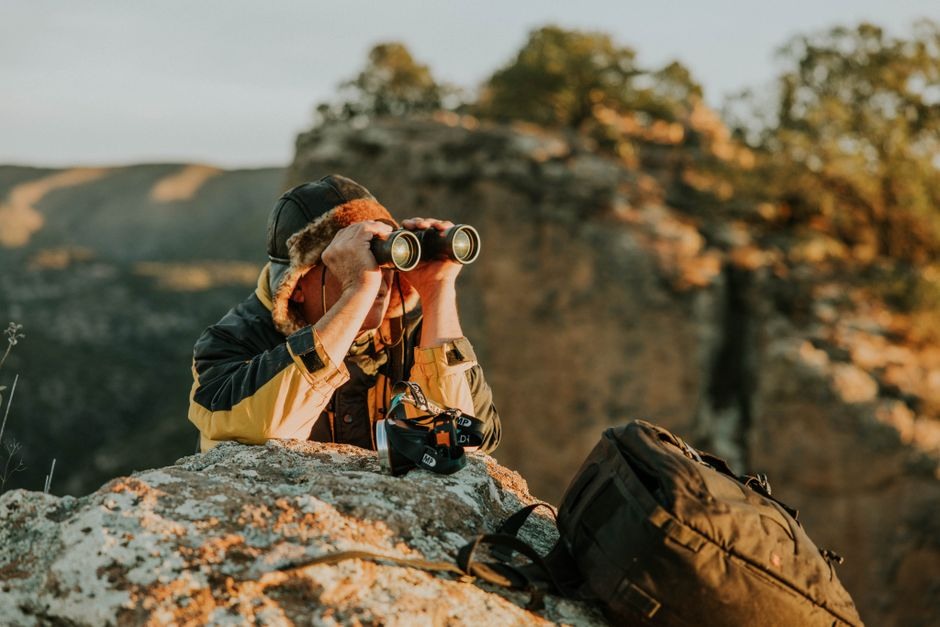 A traveler uses binoculars from a rocky viewpoint in warm evening light.