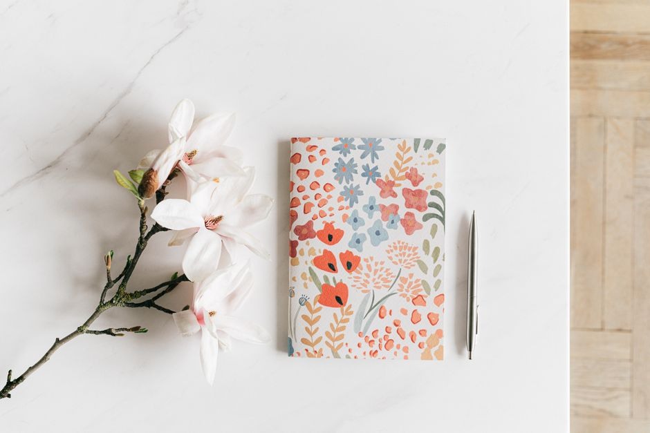 A floral notebook and pen beside pale flowers on a light surface.